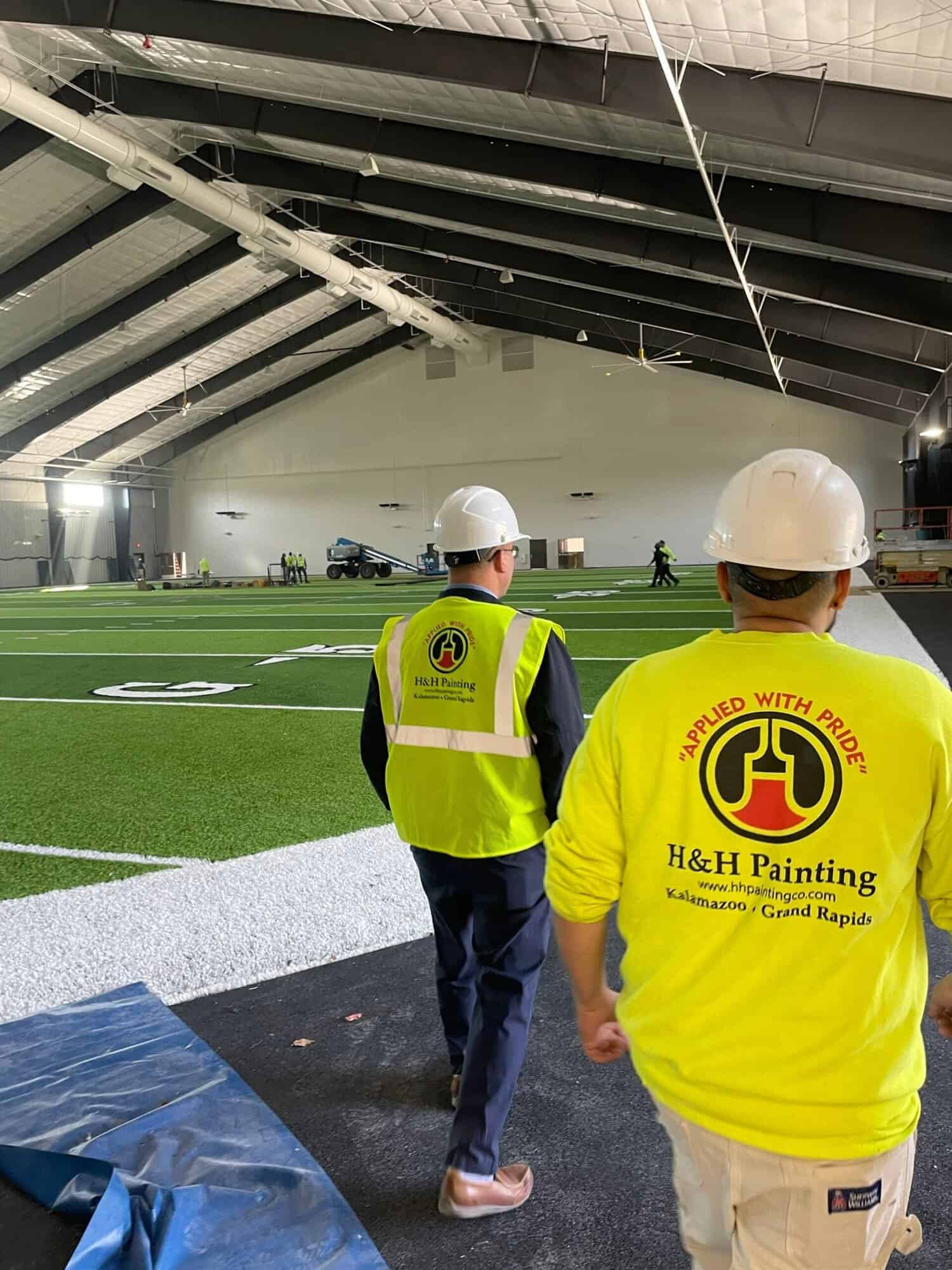 H&H Painting crew in safety gear inspecting white-painted commercial indoor sports facility with artificial turf field and