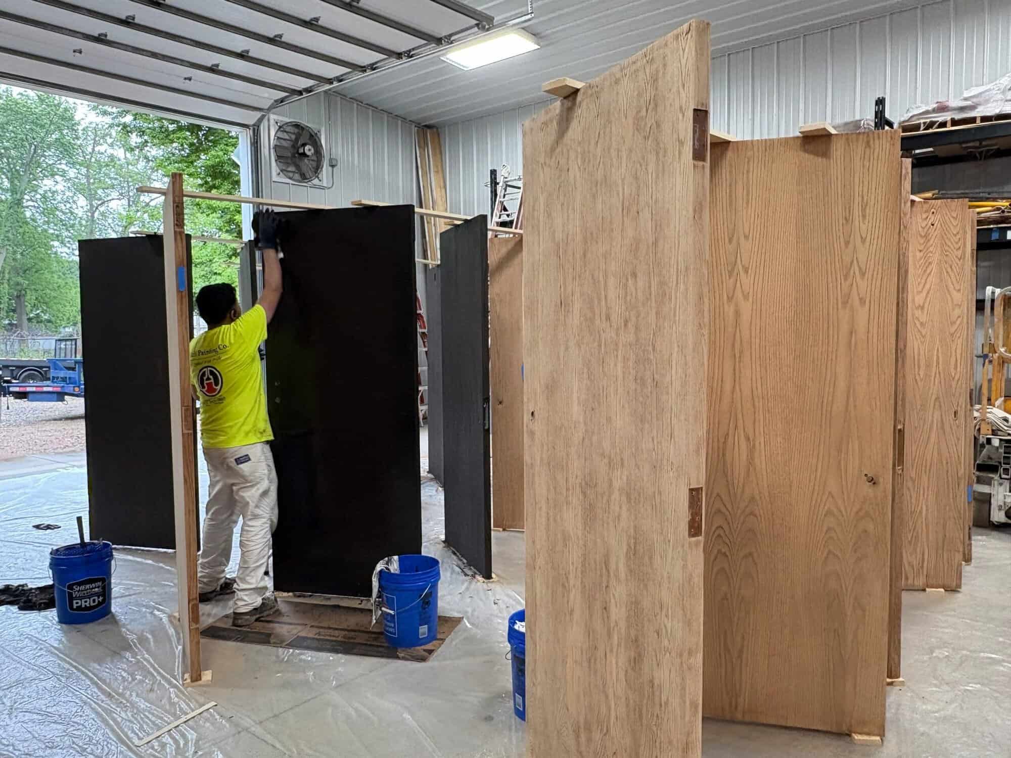 Professional Interior Door Painting Services A painter applies dark paint to a wooden door panel in a workshop, surrounded by other doors ready for refinishing.