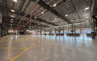 Gray polished concrete warehouse floor with yellow line markings, red structural columns, and metal truss ceiling. Commercial Epoxy Floor Coating Cost in Michigan