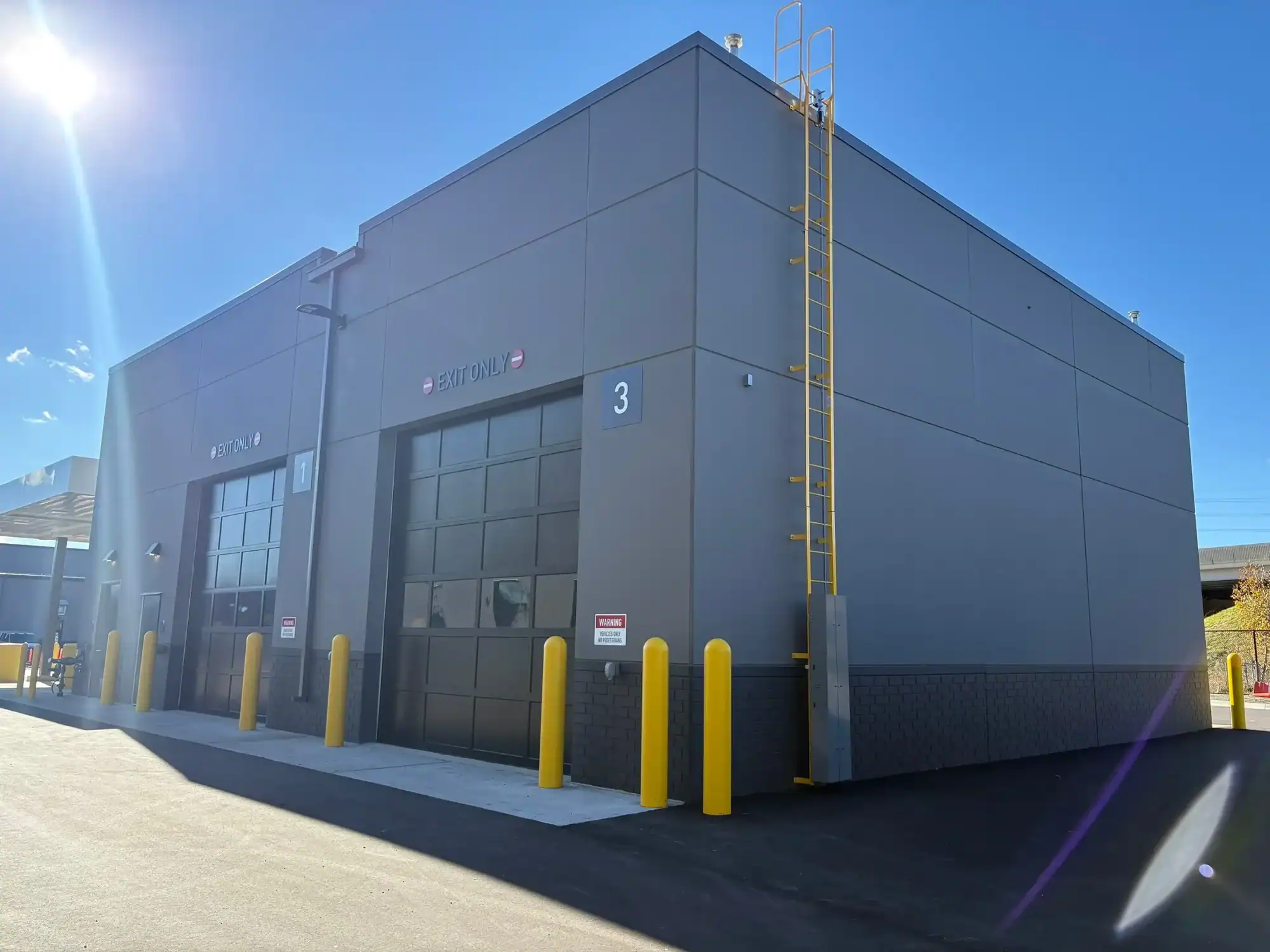 Gray Commercial Building Exterior Gray-painted commercial building exterior with black overhead doors, yellow bollards, and clear blue sky.
